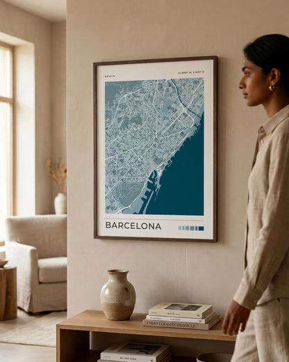 A framed Barcelona city map print hangs on a beige wall in a modern living room, with a woman standing beside it and a wooden console table below holding books and a vase.