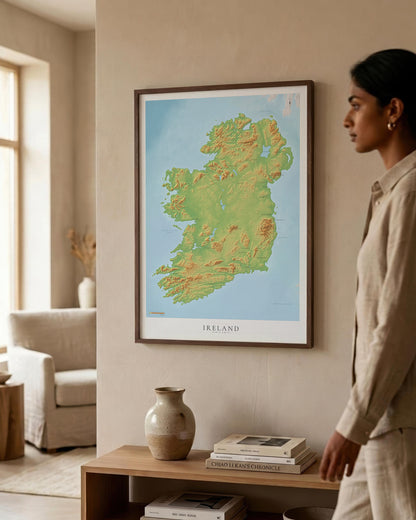 A framed topographic map of Ireland hangs on a beige wall in a modern living room, with a woman in a beige linen outfit standing to the right and a wooden console table below with books and a ceramic vase.