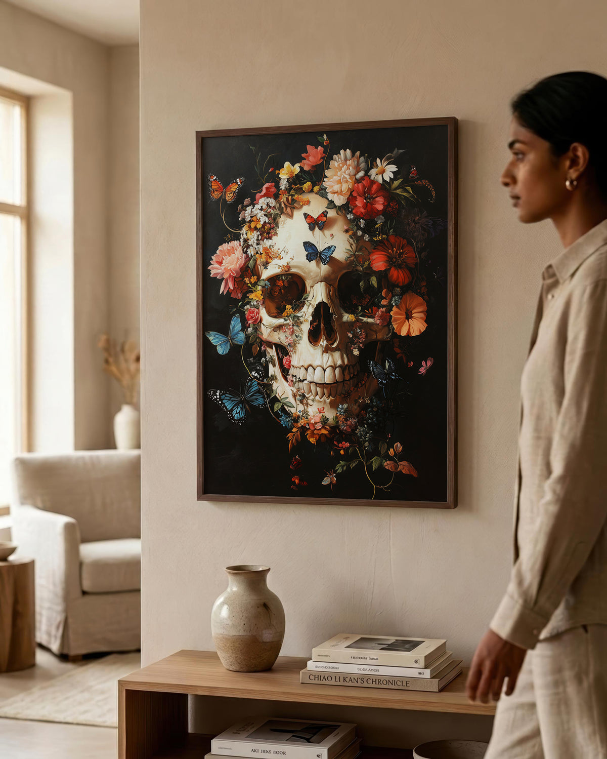 A woman in a beige linen outfit stands beside a framed wall art print showing a detailed skull surrounded by colorful flowers and blue butterflies on a dark background, displayed in a warm, modern living room setting.