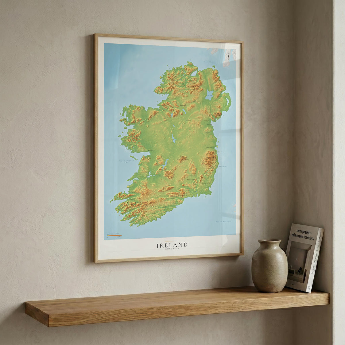 A framed topographical map poster of Ireland hangs on a textured beige wall above a wooden floating shelf. The print features green and yellow relief shading on a light blue background, with 'IRELAND' printed at the bottom.