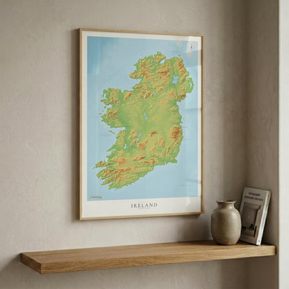 A framed topographical map poster of Ireland hangs on a textured beige wall above a wooden floating shelf. The print features green and yellow relief shading on a light blue background, with 'IRELAND' printed at the bottom.
