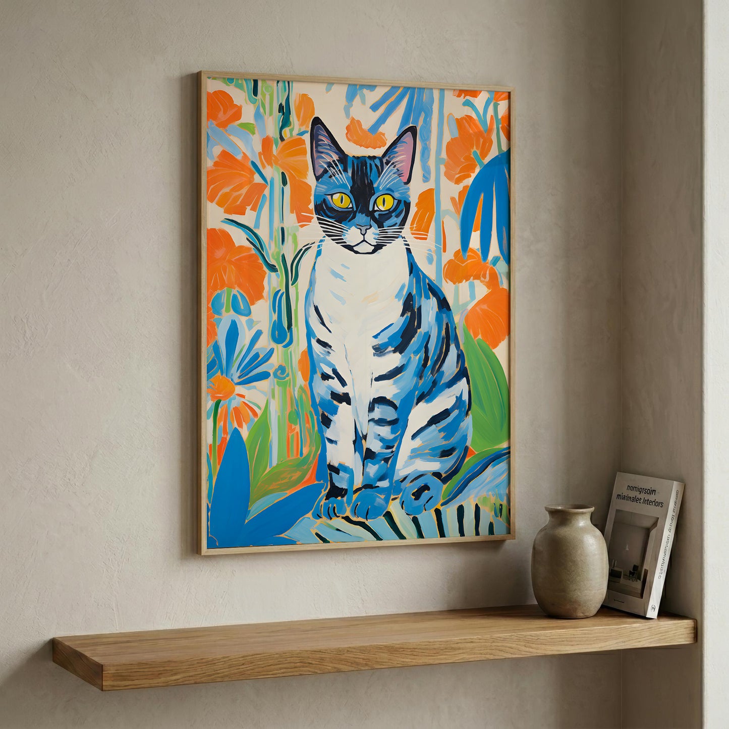 A framed colorful painting of a blue and white cat surrounded by orange flowers, displayed on a beige wall above a wooden shelf with a small vase and framed print.