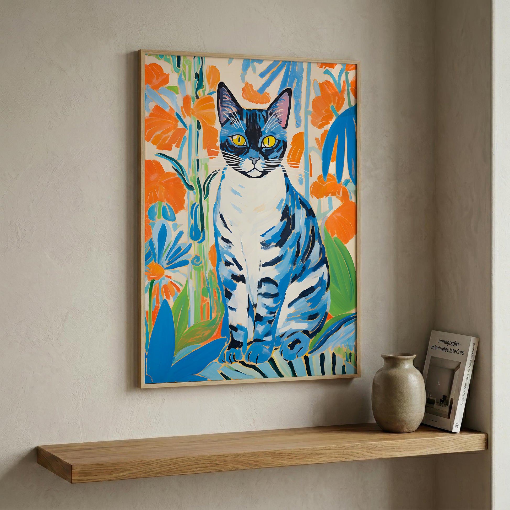 A framed colorful painting of a blue and white cat surrounded by orange flowers, displayed on a beige wall above a wooden shelf with a small vase and framed print.