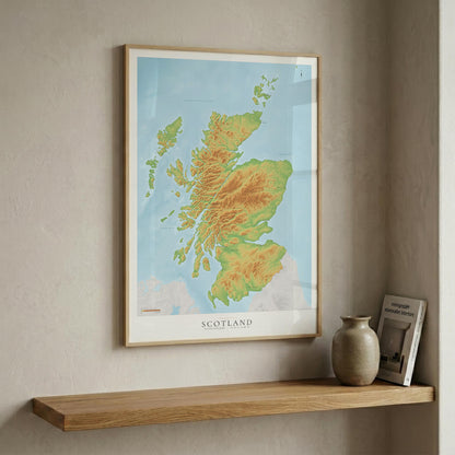A framed topographic map of Scotland, featuring green and yellow terrain shading against a light blue background, hanging on a textured beige wall above a wooden shelf with a ceramic vase and a book.