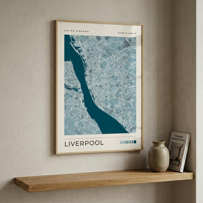 A framed map poster of Liverpool, United Kingdom, hanging on a textured beige wall above a wooden shelf with a ceramic vase and a book.