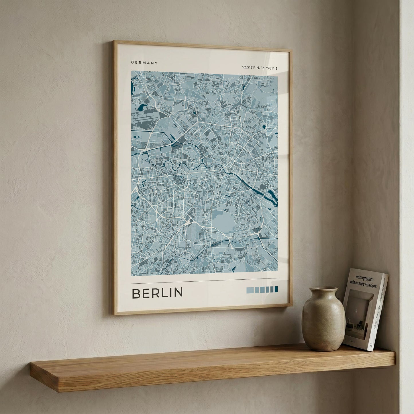 A framed city map print of Berlin, Germany, in light blue and white tones with a light wood frame, displayed on a beige wall above a wooden shelf with a ceramic vase and a book.