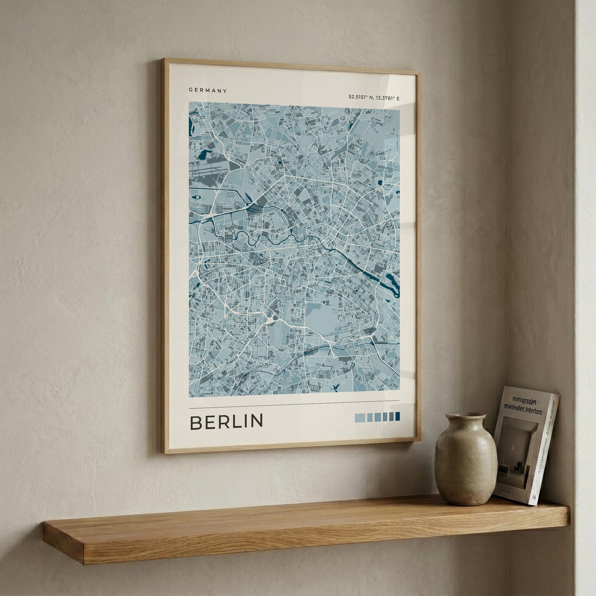 A framed city map print of Berlin, Germany, in light blue and white tones with a light wood frame, displayed on a beige wall above a wooden shelf with a ceramic vase and a book.