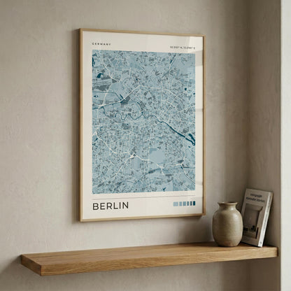A framed city map print of Berlin, Germany, in light blue and white tones with a light wood frame, displayed on a beige wall above a wooden shelf with a ceramic vase and a book.