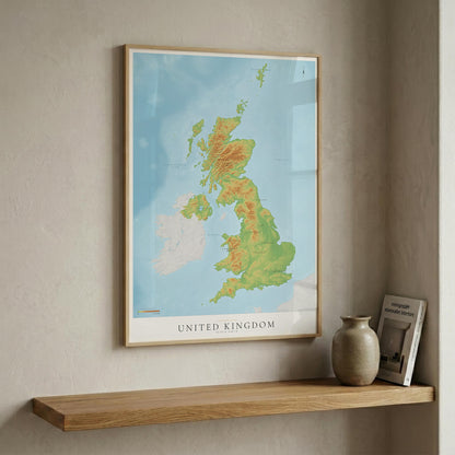 A framed topographical map poster of the United Kingdom hangs on a textured beige wall above a wooden floating shelf. The map features green, yellow, and orange terrain shading on a light blue ocean background, enclosed in a light wood frame.