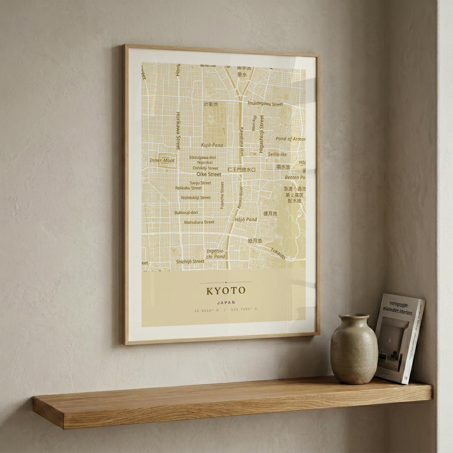 A framed vintage-style map print of Kyoto, Japan, hanging on a textured beige wall above a wooden floating shelf with a ceramic vase and a small book.