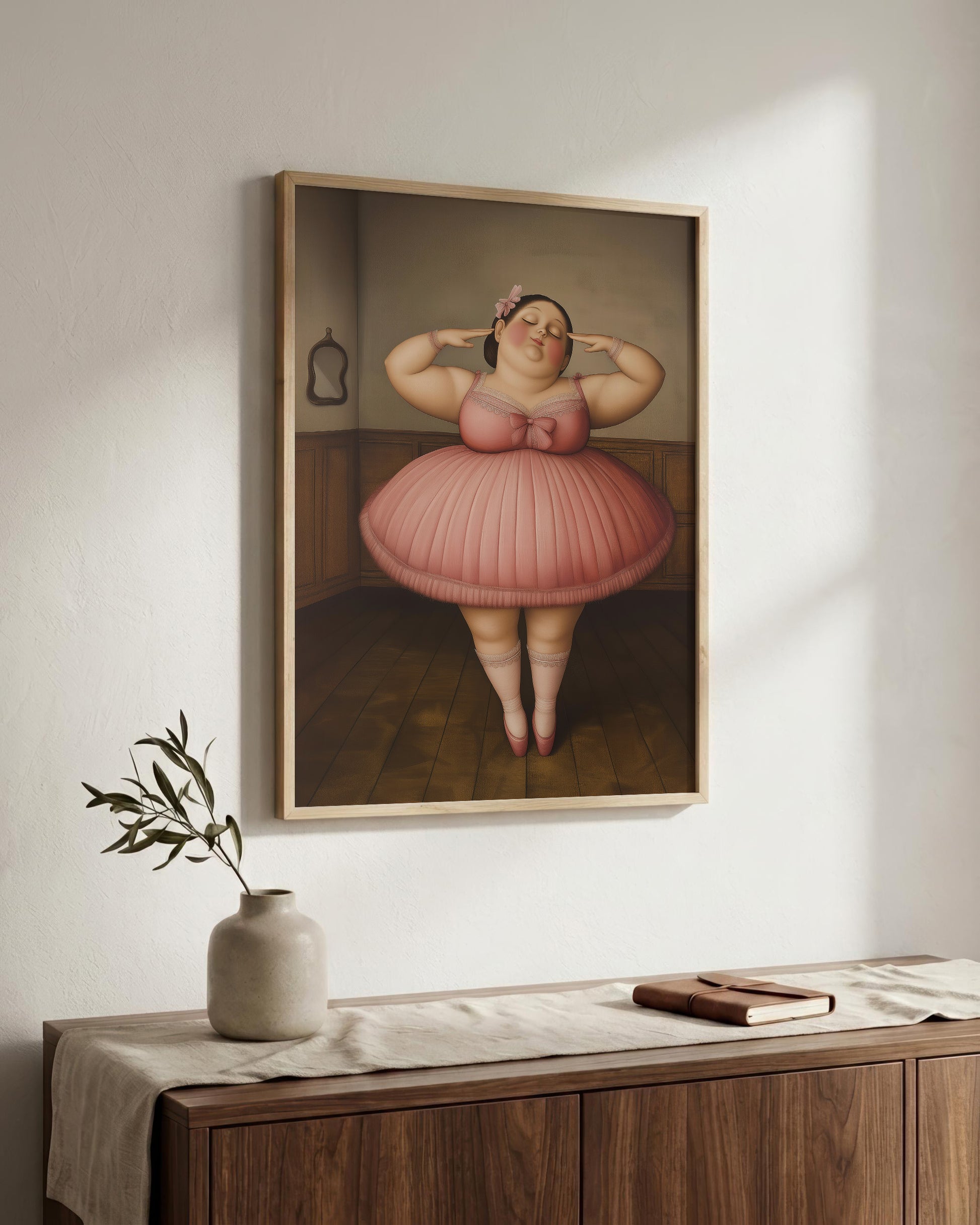 A framed art print depicting a cartoon-style figure in a pink tutu standing in a room, displayed on a white wall above a wooden console table with a vase and book.