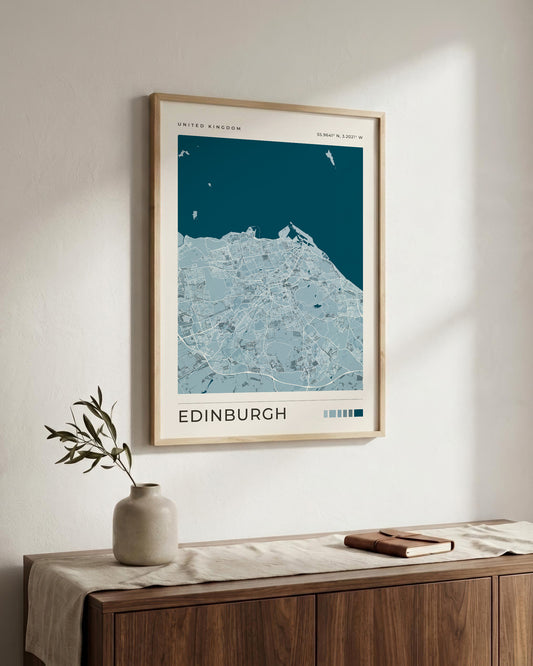 A framed Edinburgh city map poster with a light wood frame, hanging on a white wall above a wooden console table with a vase and a book.