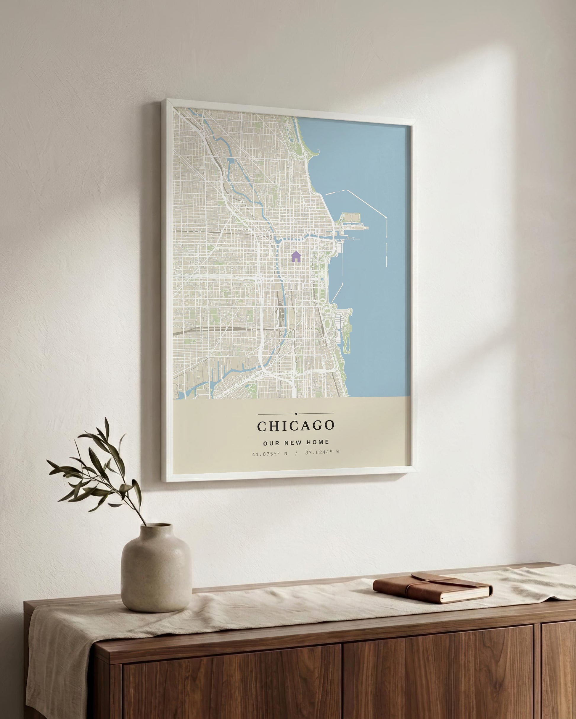 A framed Chicago city map wall print hangs on a light neutral wall above a wooden console table with a vase and book. The print features a pale blue lake area, cream and beige street grid, and the word CHICAGO at the bottom.