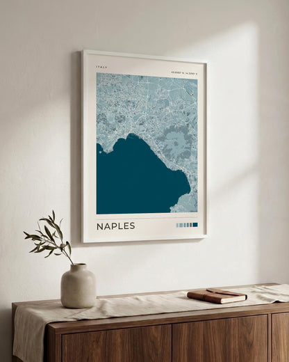 A framed Naples city map print hangs on a light wall above a wooden console table with a vase and book. The print features a dark teal coastline and city silhouette with pale blue street details and the word NAPLES at the bottom.