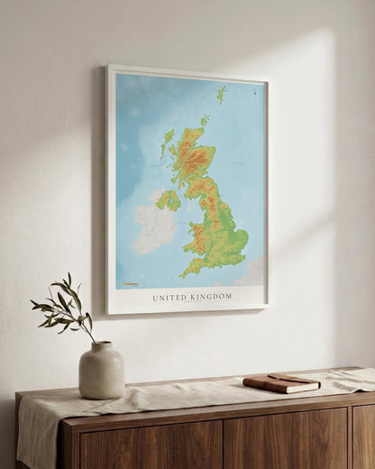A framed topographical map poster of the United Kingdom hanging on a light-colored wall above a wooden console table with a vase and a book.