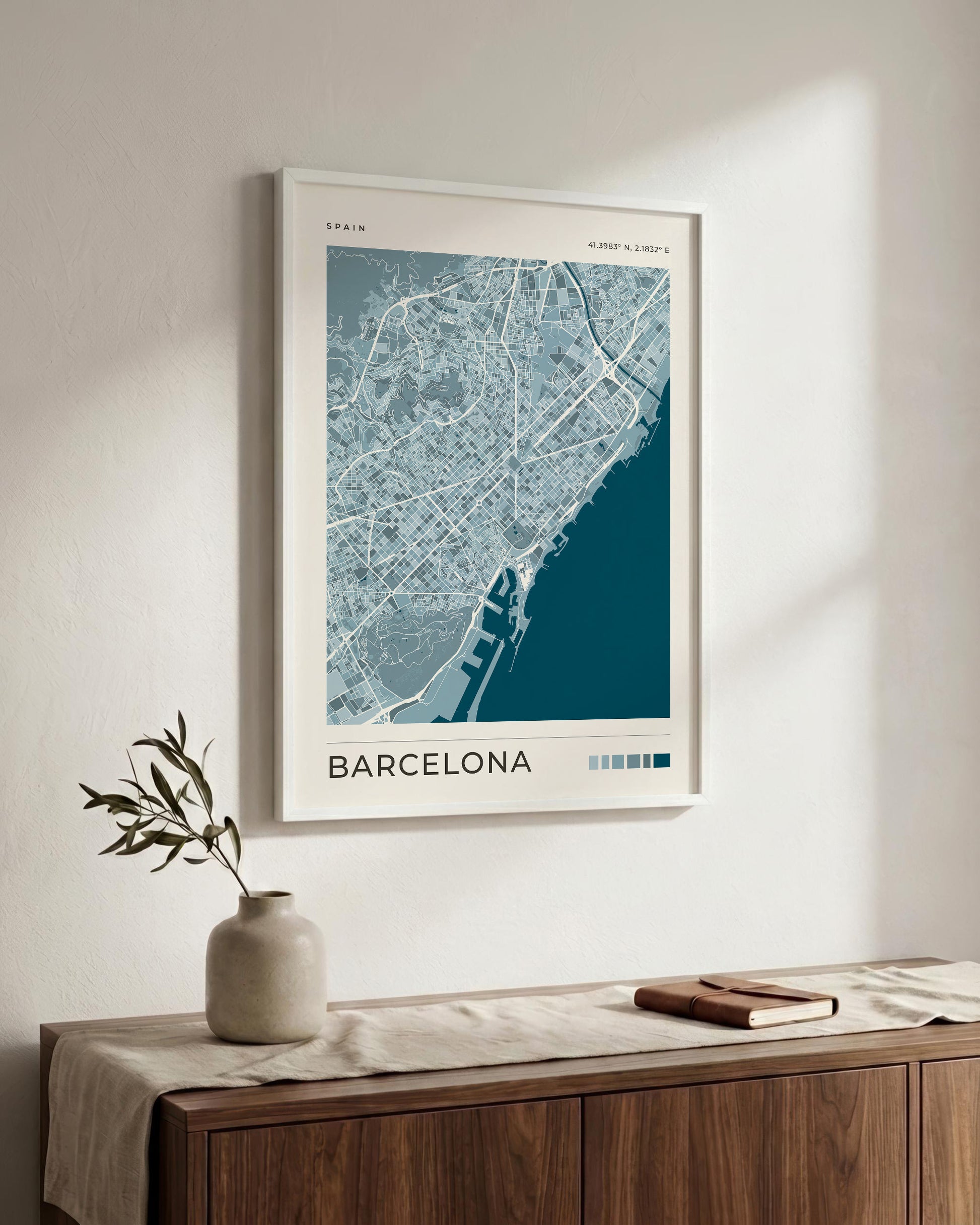 A framed Barcelona city map print hangs on a light beige wall above a wooden sideboard with a vase and book. The poster shows a blue and white street map with the word BARCELONA at the bottom.