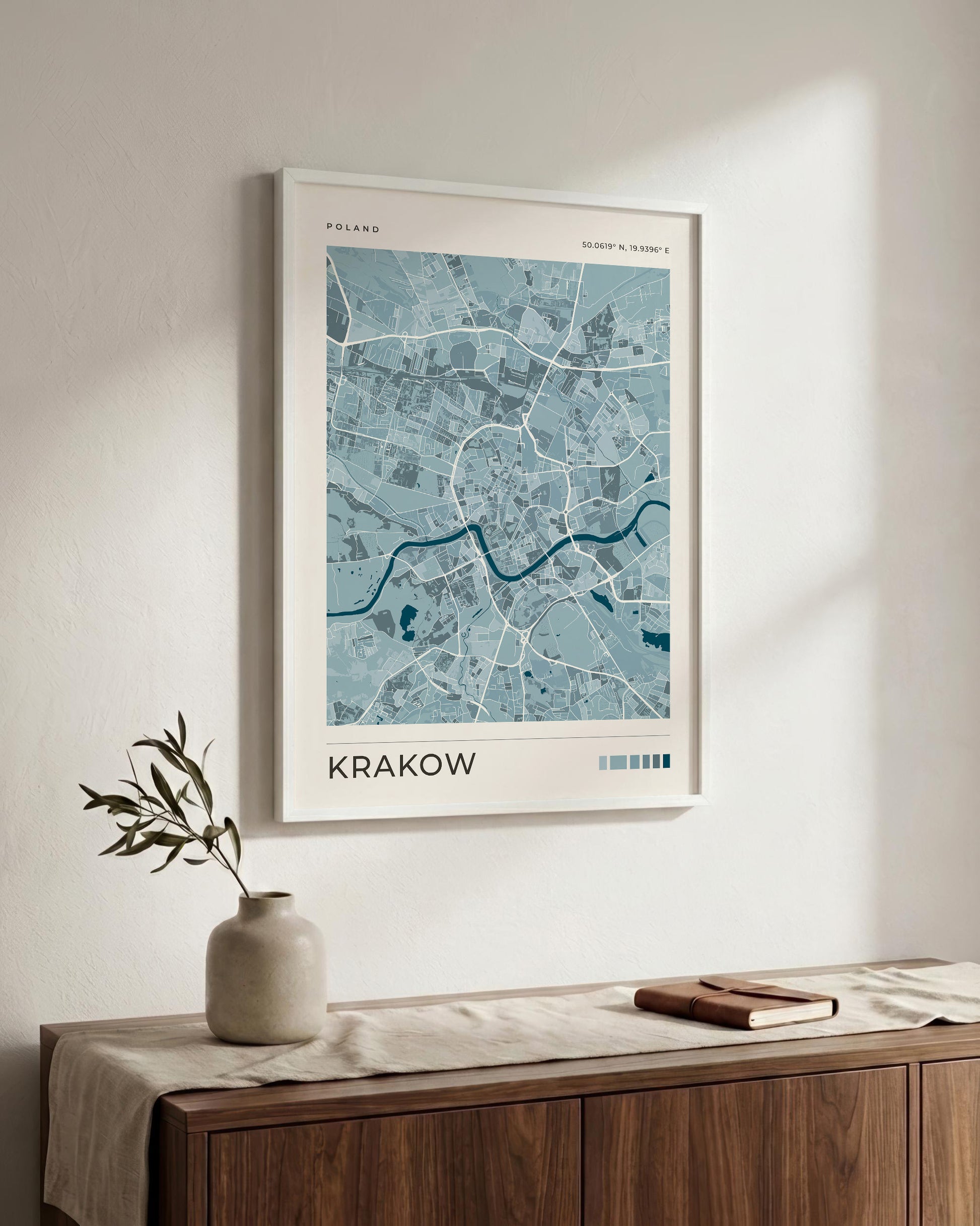 A framed city map print of Krakow, Poland, featuring a light blue and teal map design with white roads and the word 'KRAKOW' at the bottom, displayed on a warm off-white wall above a wooden sideboard with a vase and a book.