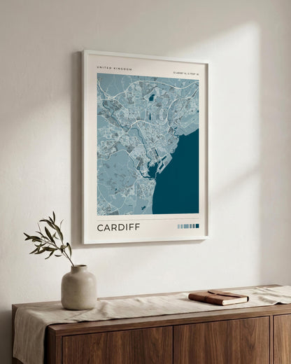 A framed city map print of Cardiff, Wales, displayed on a light beige wall above a wooden sideboard with a vase and book. The print features a light blue and white map with a dark teal coastal area and the word 'CARDIFF' at the bottom.