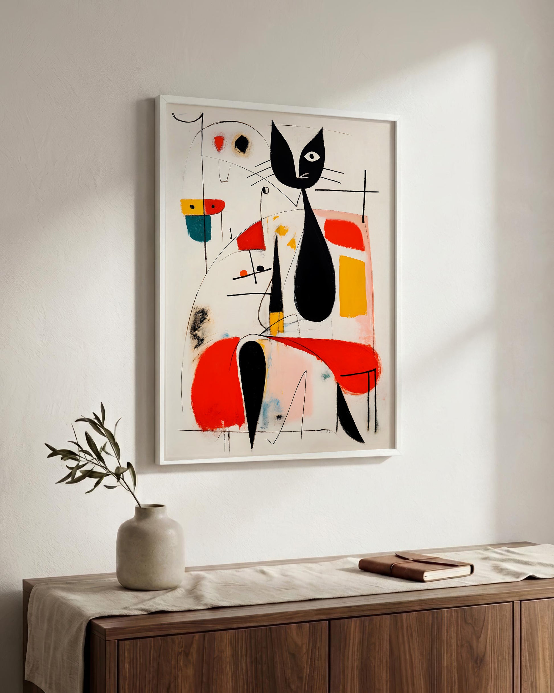 A framed abstract art print featuring a stylized black cat with red, yellow, and blue geometric shapes, displayed on a white wall above a wooden console table with a vase and book.