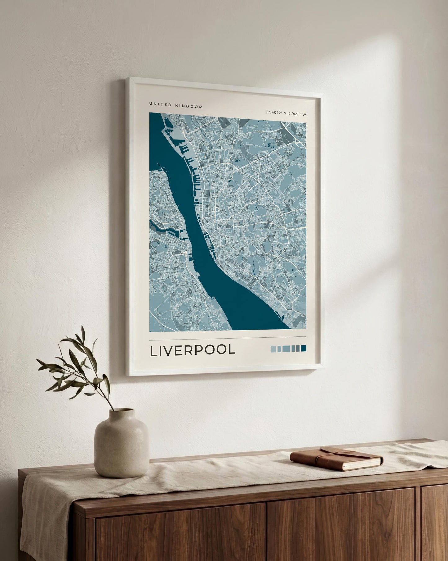 A framed map print of Liverpool, United Kingdom, featuring a blue and white street layout with a dark teal river, displayed on a light wall above a wooden console table with a vase and a book.