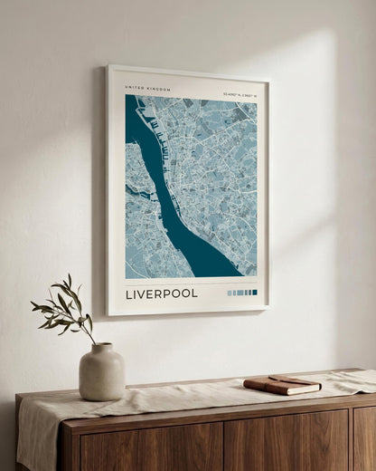 A framed map print of Liverpool, United Kingdom, featuring a blue and white street layout with a dark teal river, displayed on a light wall above a wooden console table with a vase and a book.