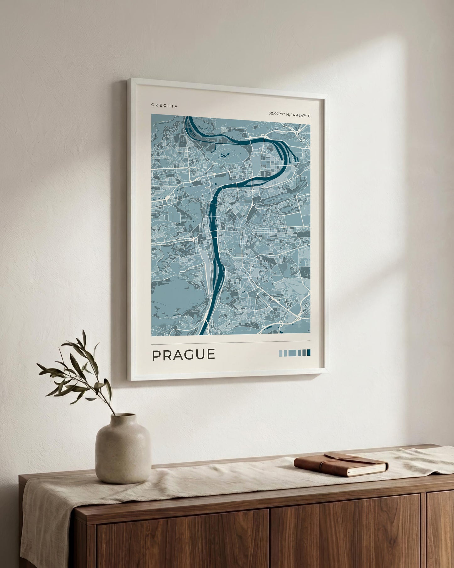 A framed art print of a blue and gray map of Prague, featuring the Vltava River, displayed on a warm off-white wall above a wooden console table with a vase and a book.