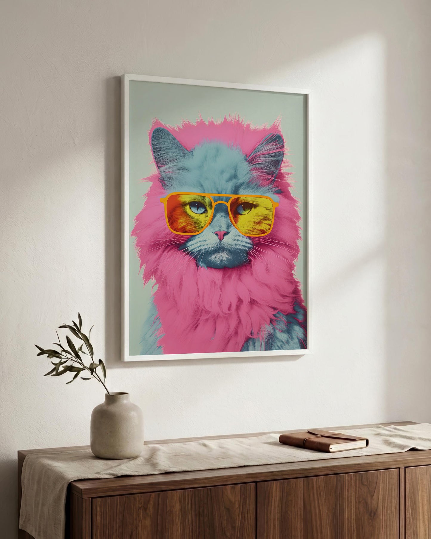 A framed pop-art style poster of a fluffy cat wearing pink and yellow sunglasses, displayed on a light wall above a wooden sideboard with a vase and book.