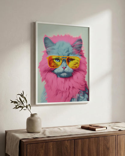 A framed pop-art style poster of a fluffy cat wearing pink and yellow sunglasses, displayed on a light wall above a wooden sideboard with a vase and book.