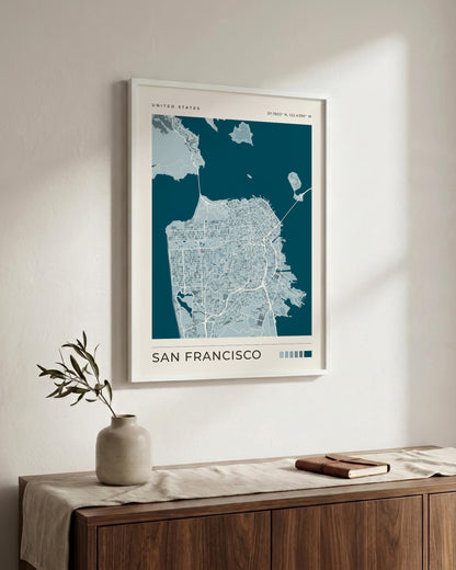 A framed San Francisco city map print hangs on a light beige wall above a wooden console table with a vase and a book. The print features a deep teal background with white street lines and the text 'UNITED STATES' and 'SAN FRANCISCO'.