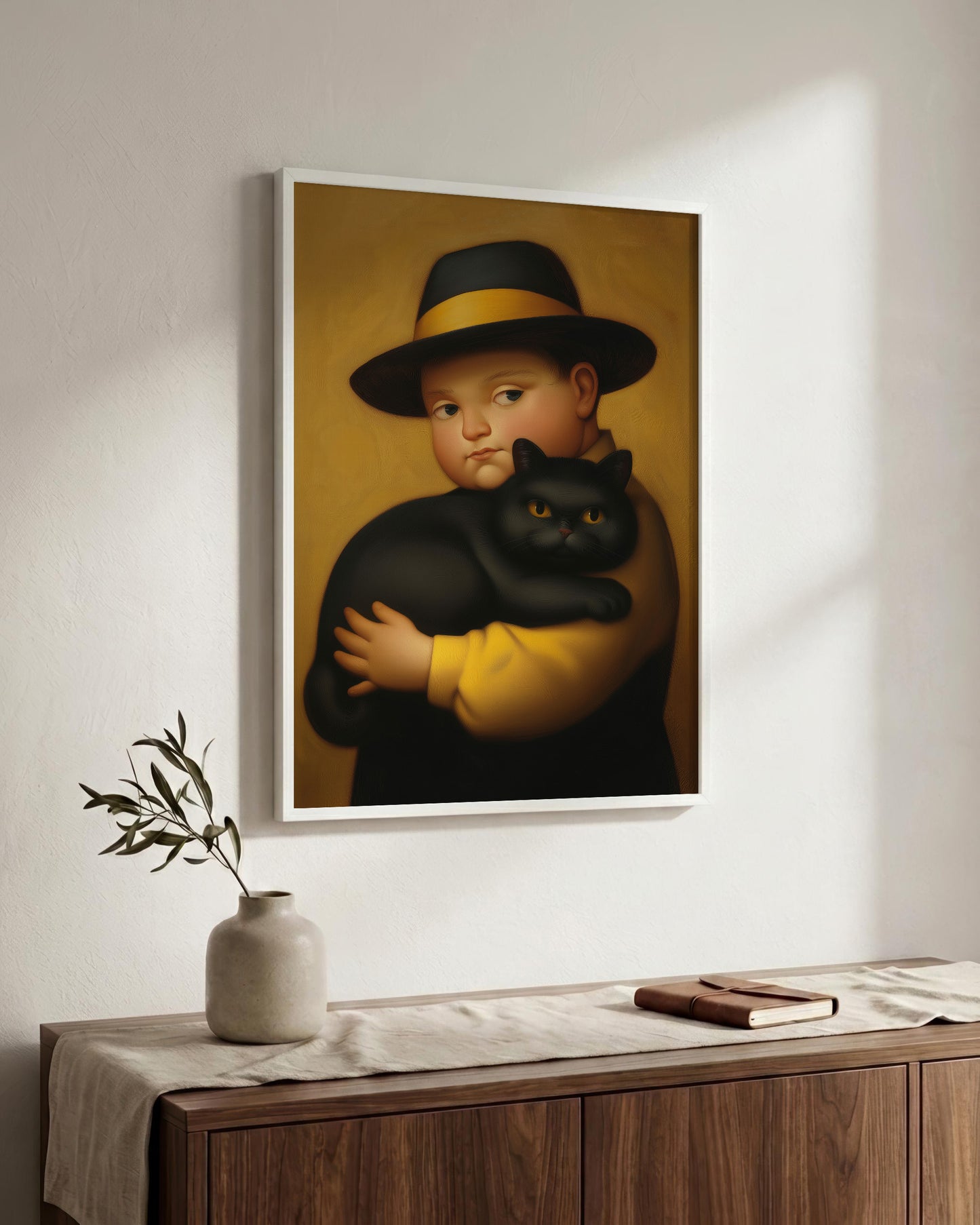A framed art print depicting a classical-style portrait of a boy in a black hat with a yellow band, holding a black cat, hung on a white wall above a wooden console table with a vase and book.