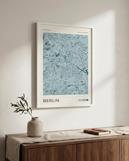 A framed art print of a light blue and white map of Berlin, Germany, hanging on a textured white wall above a wooden sideboard with a vase and a book.
