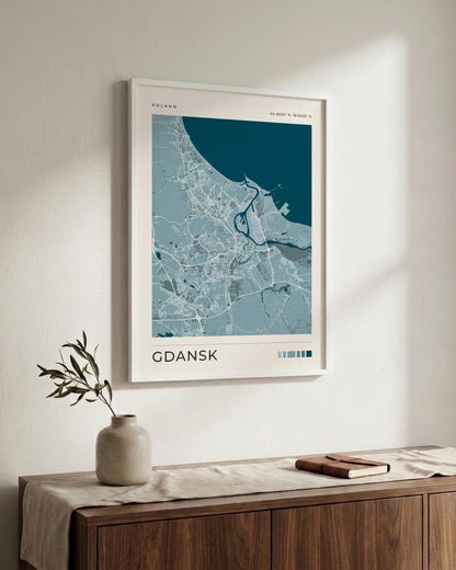 A framed city map print of Gdansk, Poland, featuring a light blue and white map design with a dark teal sea area, hanging on a warm off-white wall above a wooden sideboard with a vase and book.