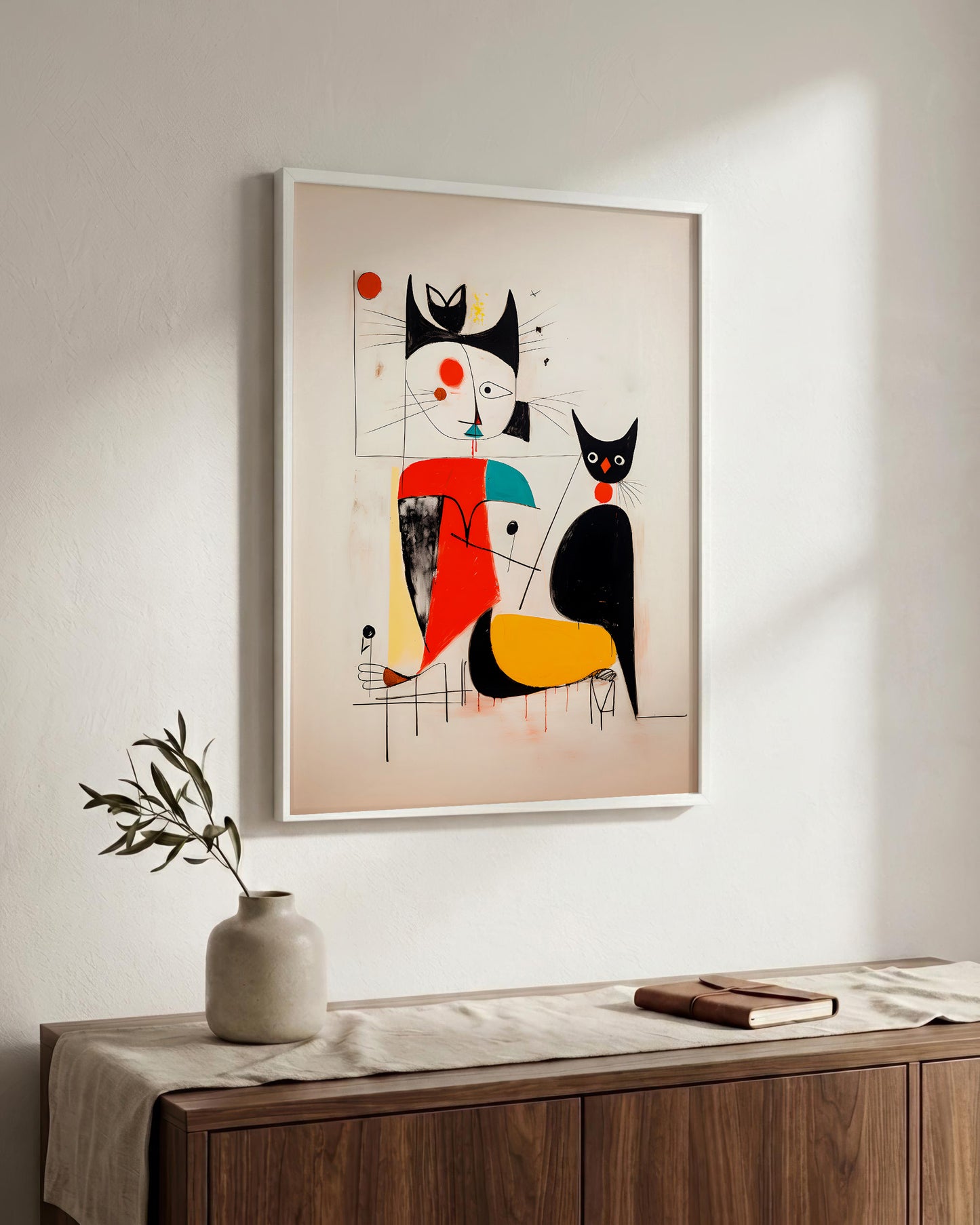 A framed abstract art print featuring stylized black and red cat-like figures on a beige background, hanging on a white wall above a wooden console table with a vase and book.