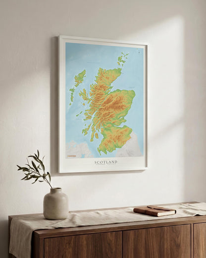 A framed Scotland topographic map print hangs on a light beige wall above a wooden sideboard with a vase and book in a styled interior setting.