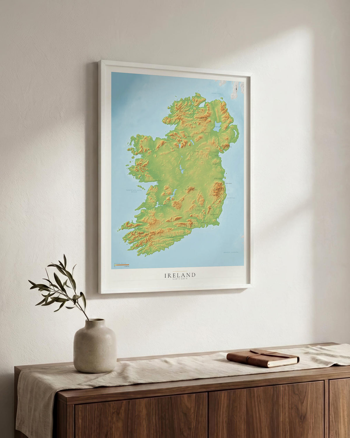 A framed topographical map print of Ireland hangs on a light wall above a wooden sideboard with a vase and book in a styled interior setting.