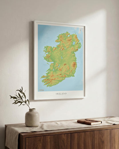A framed topographical map print of Ireland hangs on a light wall above a wooden sideboard with a vase and book in a styled interior setting.