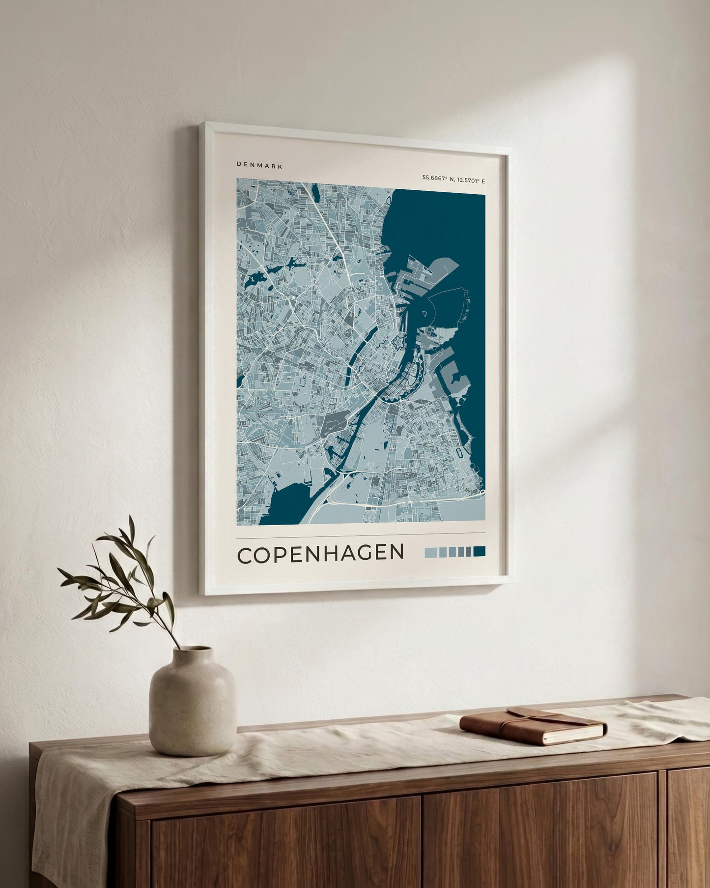 A framed Copenhagen city map art print hangs on a light beige wall above a wooden sideboard with a vase and book in a styled interior setting.