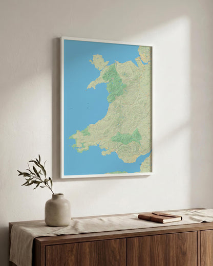 A framed map print of Wales hangs on a light beige wall above a wooden console table with a vase and a book. The print features a light blue ocean and a beige and green landmass.