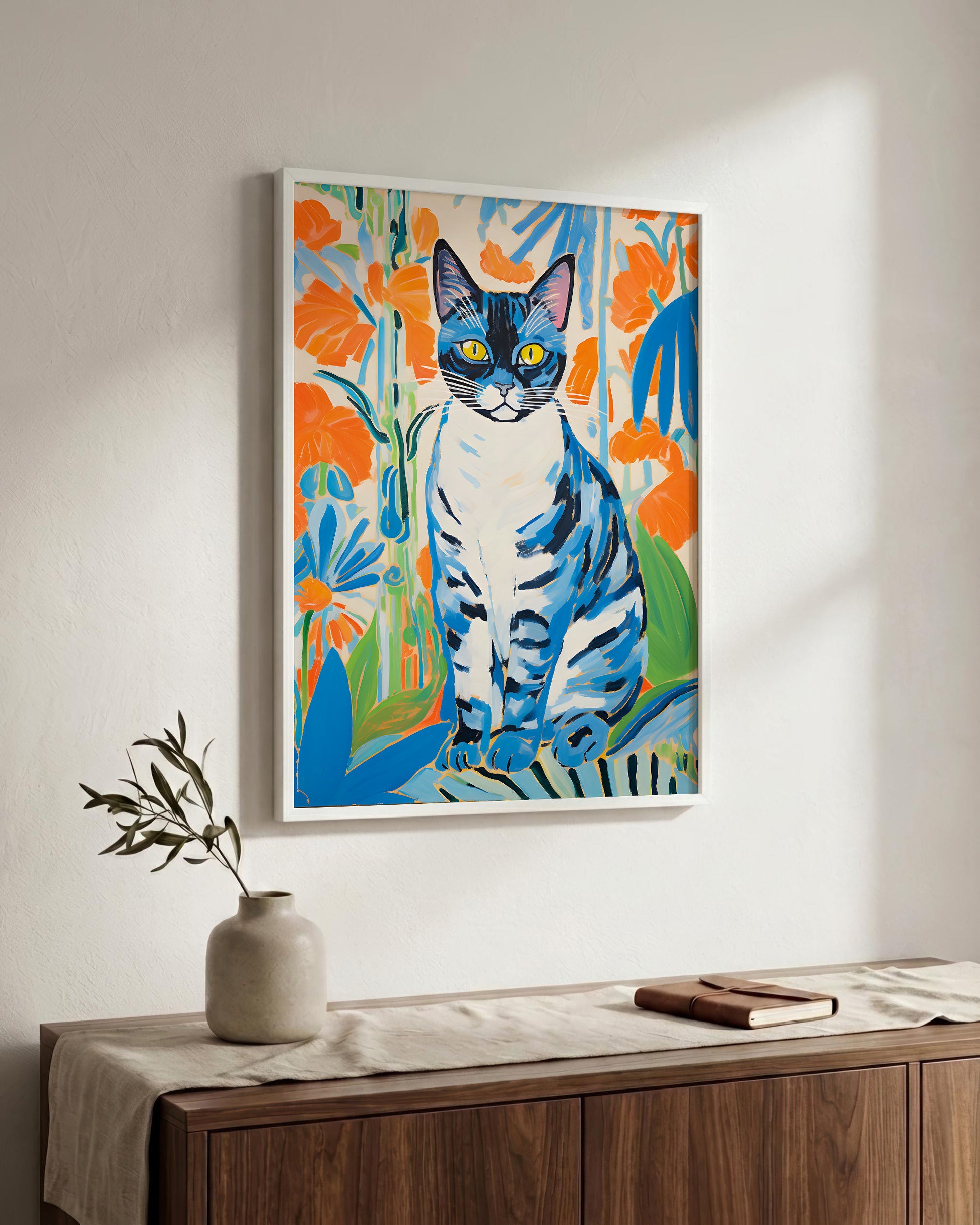 A framed colorful art print of a blue and white cat surrounded by orange flowers, hanging on a white wall above a wooden sideboard with a vase and book.