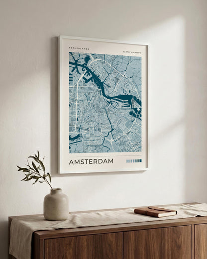 A framed Amsterdam city map print hangs on a light beige wall above a wooden sideboard with a vase and a book. The poster has a white border and features blue street lines and waterways, with the word 'AMSTERDAM' printed at the bottom.
