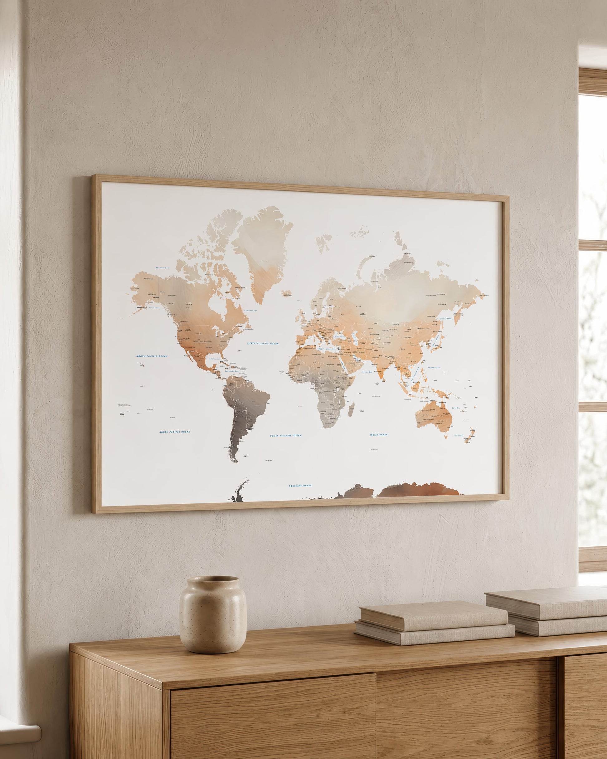 A framed world map art print with a light wood frame, displayed on a textured beige wall above a wooden console table with books and a ceramic vase.
