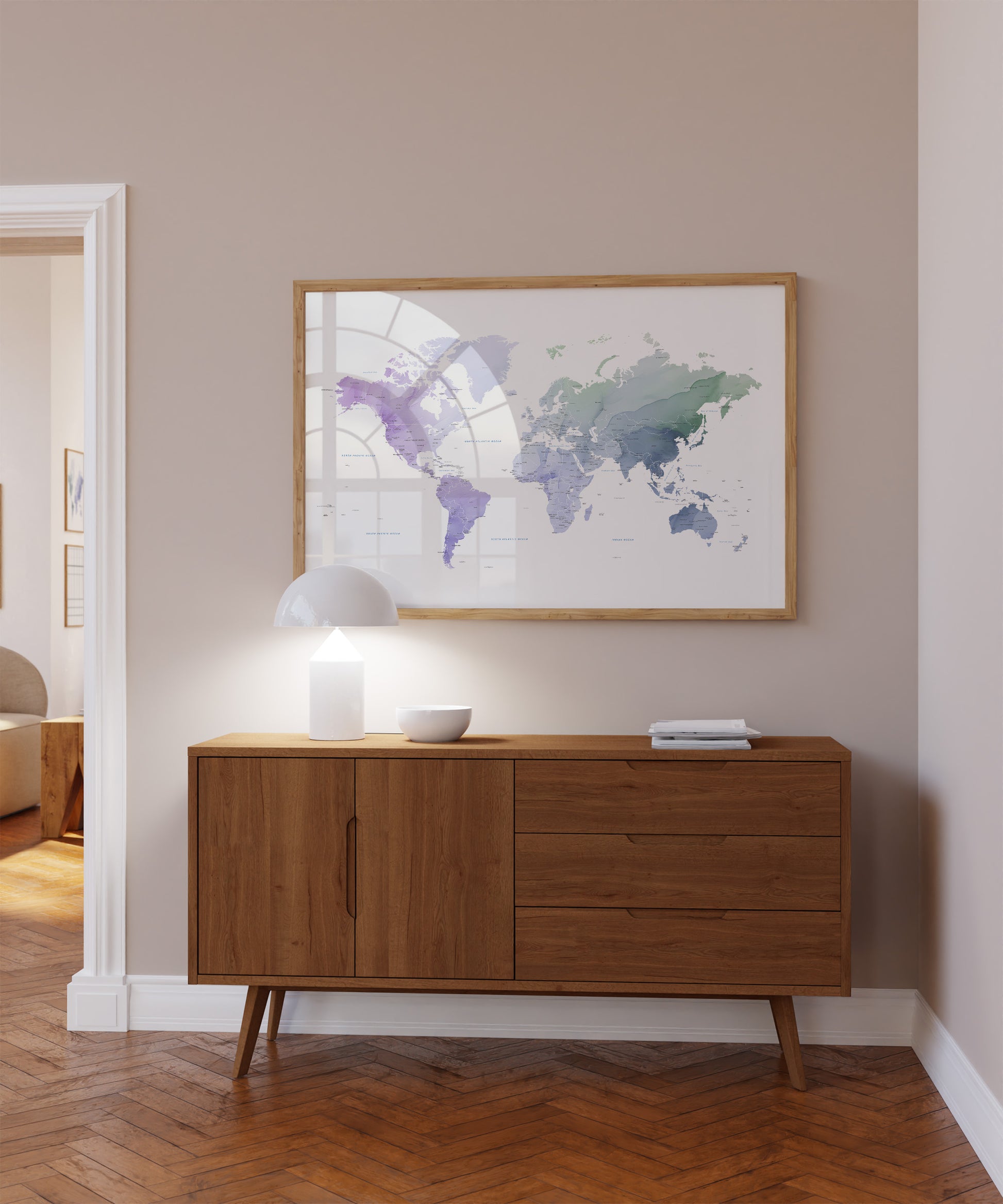 A framed world map print with watercolor-style purple and green landmasses hangs on a beige wall above a mid-century modern wooden sideboard with a lit lamp and small bowl in a styled room.
