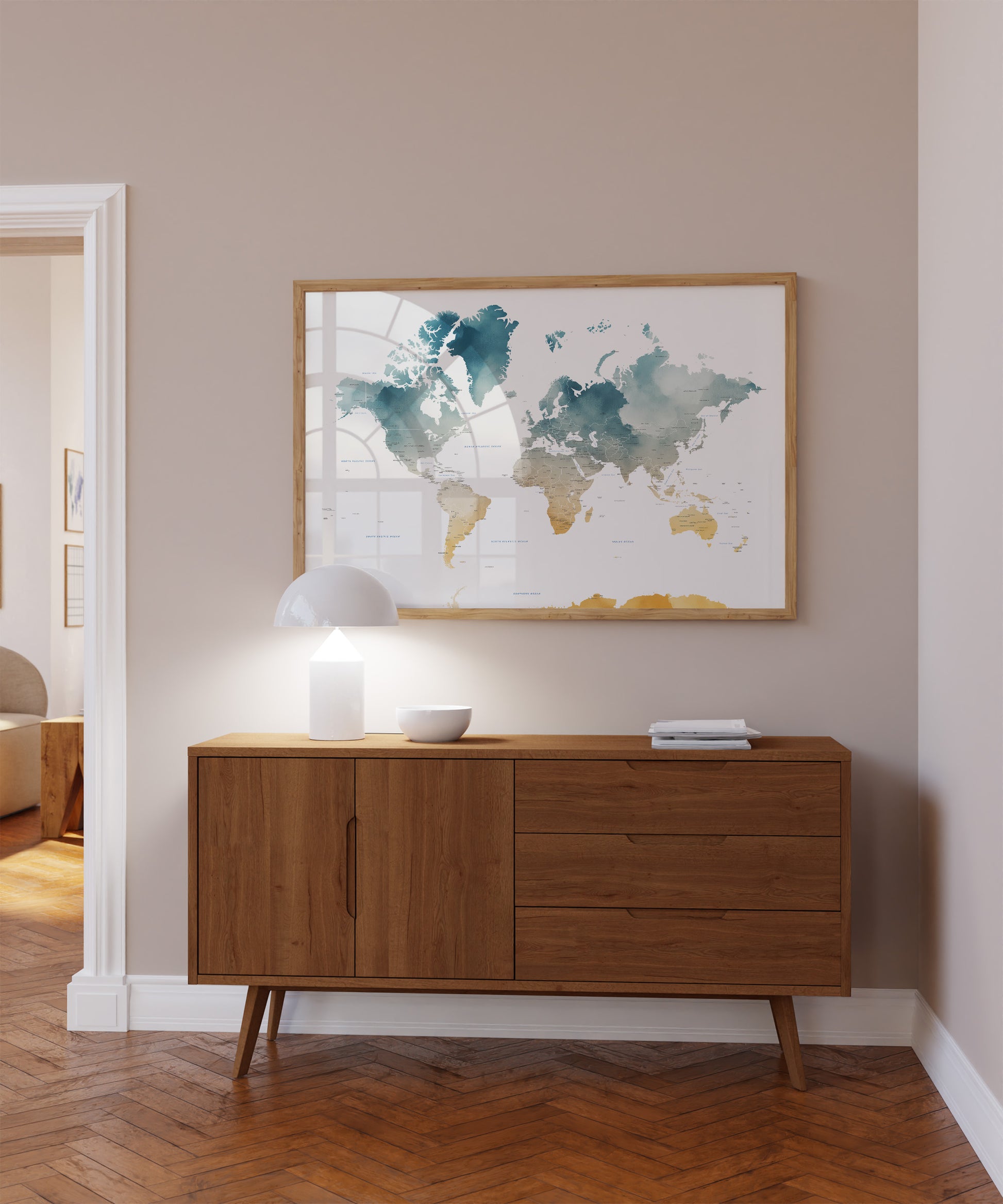 A framed world map art print with watercolor-style continents in teal, blue, and gold tones, hanging on a beige wall above a mid-century modern wooden sideboard with a lit lamp and small bowls.