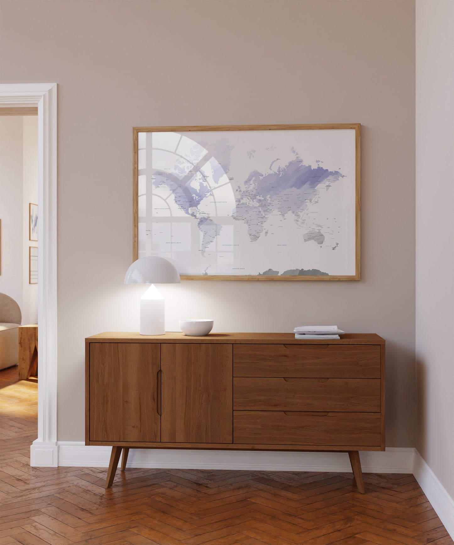 A framed world map print with a light wood frame hangs on a beige wall above a mid-century modern wooden sideboard. The room features warm wood herringbone flooring, a white dome lamp, and a small white bowl on the cabinet.