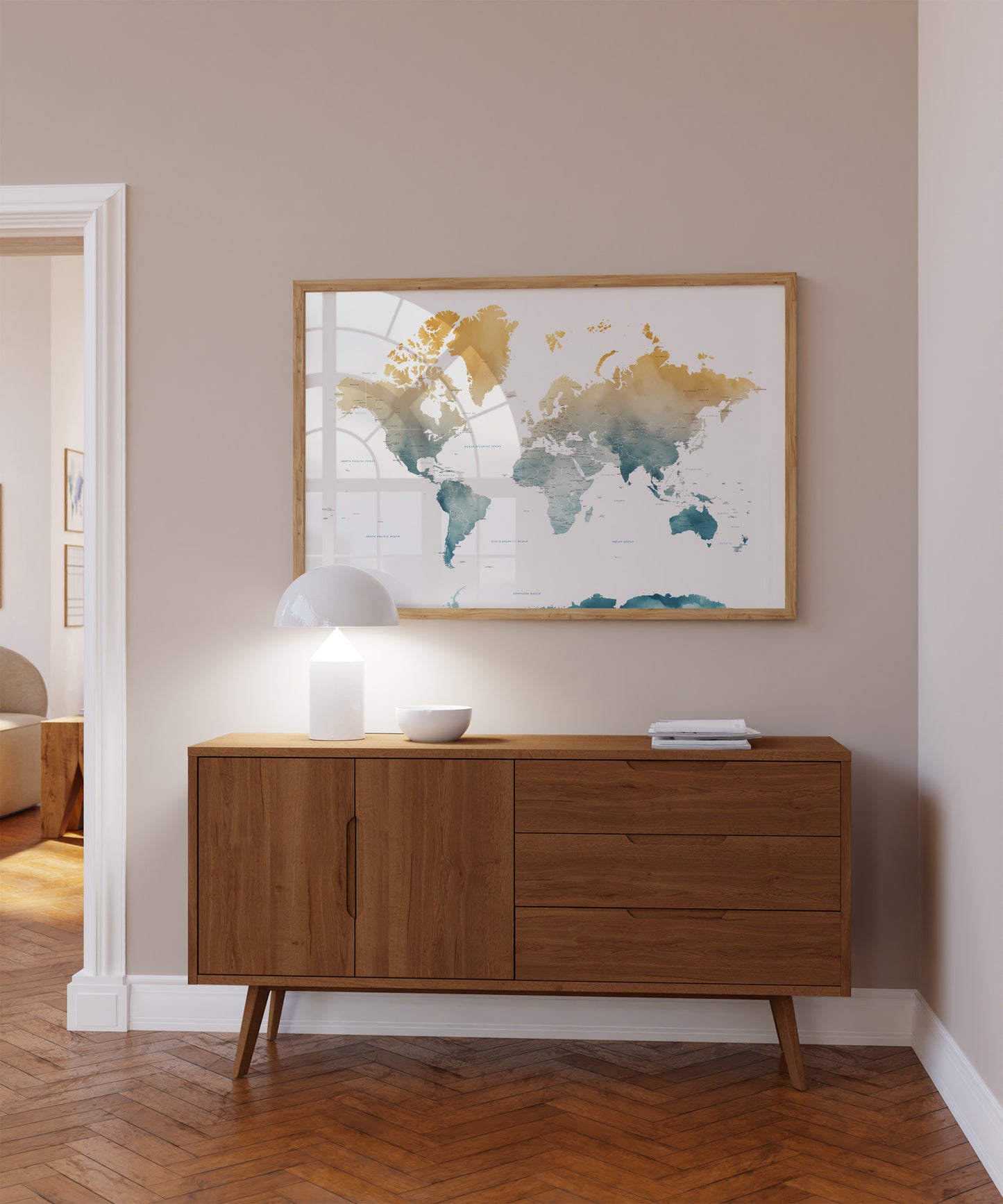 A framed world map art print with watercolor-style continents in gold, teal, and blue tones, displayed on a beige wall above a mid-century modern wooden sideboard with a lit lamp and small decor items.