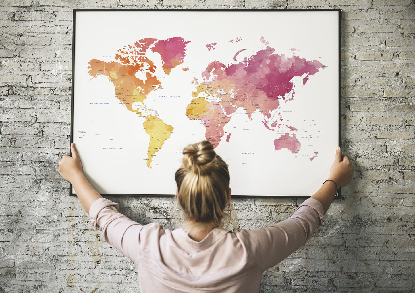 A woman with her hair in a bun hangs a large framed world map on a gray brick wall. The map features a watercolor design with continents in shades of pink, orange, and yellow on a white background.