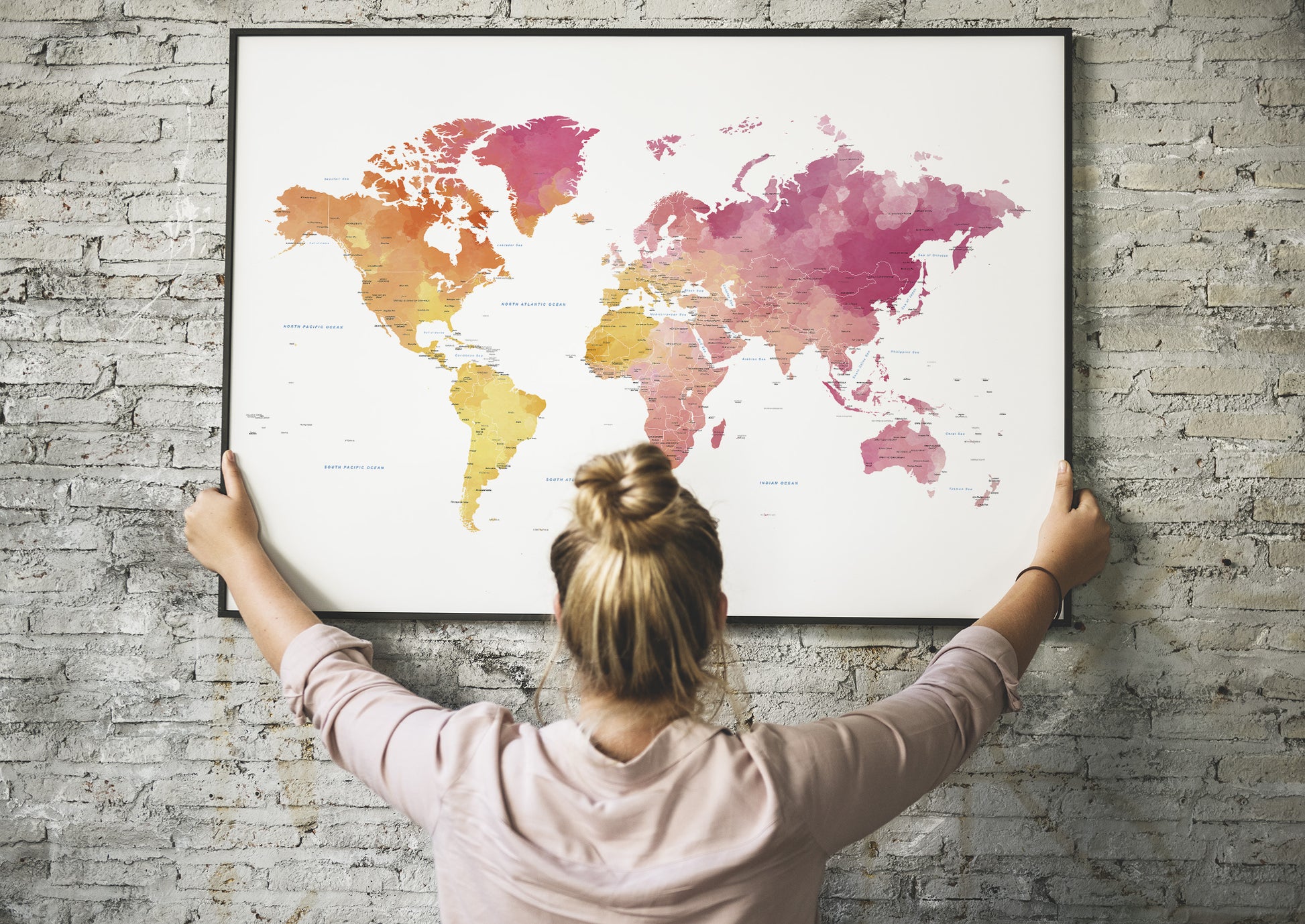 A woman with her hair in a bun hangs a large framed world map on a gray brick wall. The map features a watercolor design with continents in shades of pink, orange, and yellow on a white background.