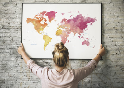 A woman with her hair in a bun hangs a large framed world map on a gray brick wall. The map features a watercolor design with continents in shades of pink, orange, and yellow on a white background.