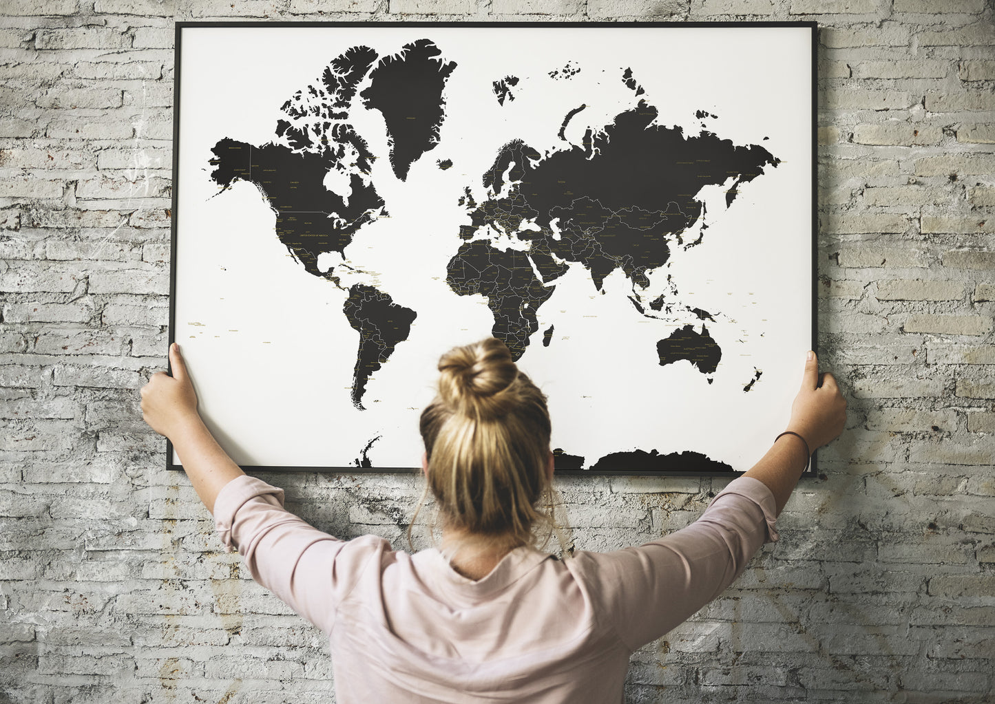 A woman with her hair in a bun hangs a large black-and-white world map poster on a light gray brick wall, viewed from behind her.