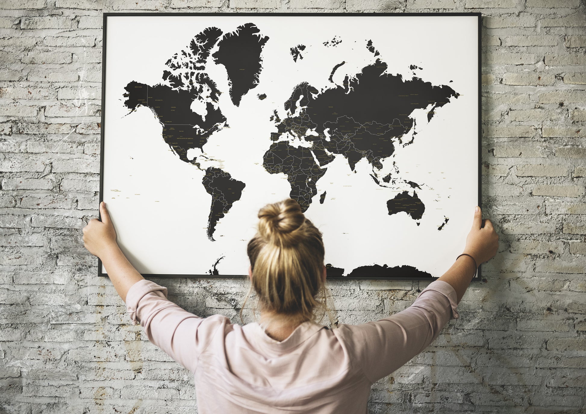 A woman with her hair in a bun hangs a large black-and-white world map poster on a light gray brick wall, viewed from behind her.